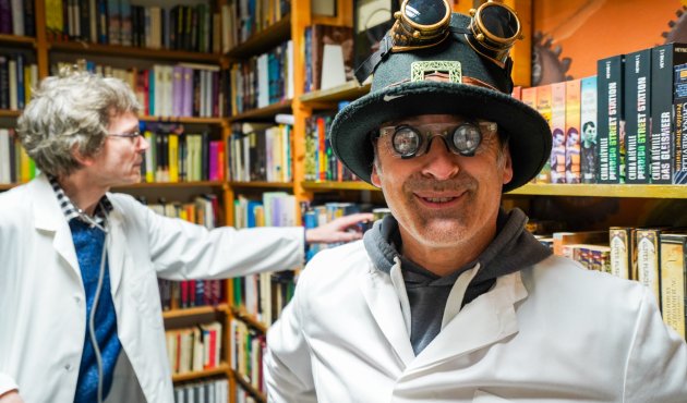 Mr. Ralf mit Steampunk-Hut und -brille, im Hintergrund Dr. Stefan an einem Bücherregal