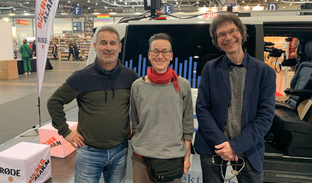 Szene in einer Messehalle: Ralf Schweikart, Josephine Mark und Stefan Hauck stehen nebeneinander vor einem Podcastbus