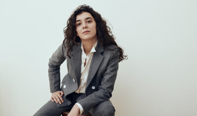 Porträtfoto von Nora Haddada, in grauem Anzug auf einem Hocker sitzend