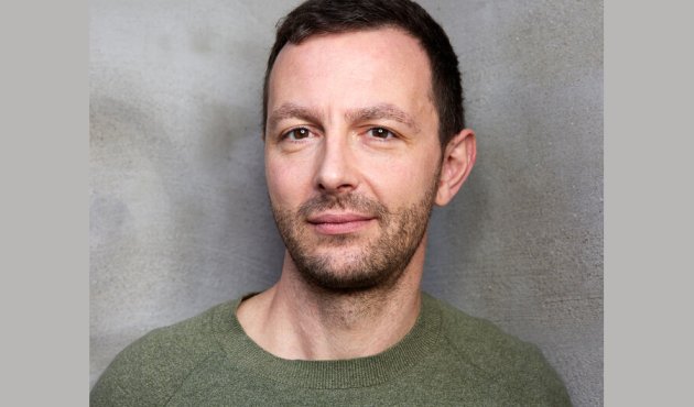 Porträt von Sebastian Guggolz mit grünem Pulli vor grauem Hintergrund