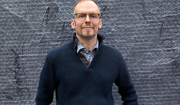 Dietmar Dath steht lächelnd vor einer grauen, strukturierten Wand