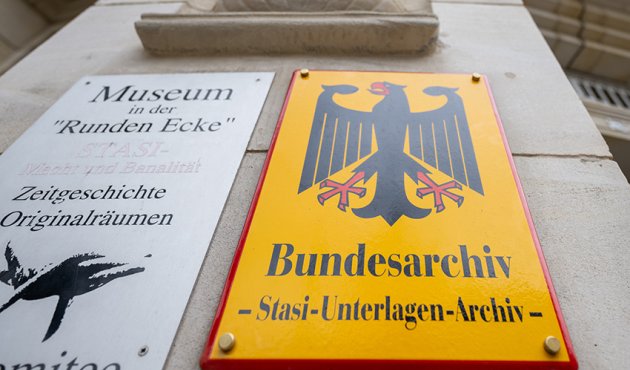 Ein Foto zeigt ein Schild mit der Aufschrift „Bundesarchiv - Stasi-Unterlagen-Archiv“ an der Außenstelle des Bundesarchivs in Leipzig. Das Schild ist gelb mit einem schwarzen Adler und befindet sich neben einem weiteren Schild, das auf ein Museum hinweist. Der Hintergrund zeigt die Fassade des Gebäudes.