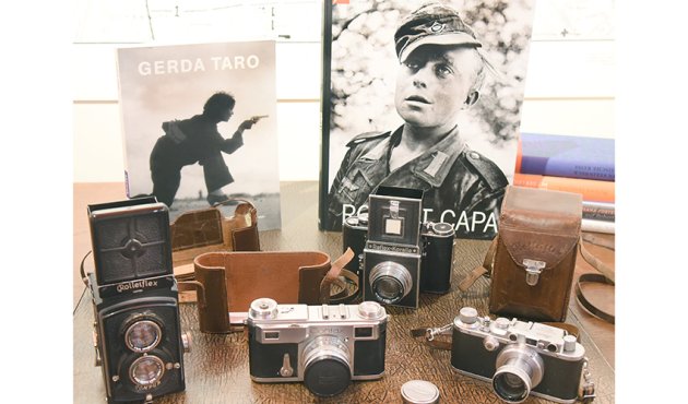 Ein Foto zeigt mehrere Kameras auf einem Schreibtisch, darunter eine Rollei, eine Contax und eine Reflex-Korelle. Im Hintergrund stehen zwei Fotobücher, eines über Gerda Taro und eines über Robert Capa. Die Kameras sind in verschiedenen Größen und Designs angeordnet, und einige sind in Lederetuis verpackt. Der Tisch hat eine strukturierte Oberfläche, und die Umgebung ist hell beleuchtet.