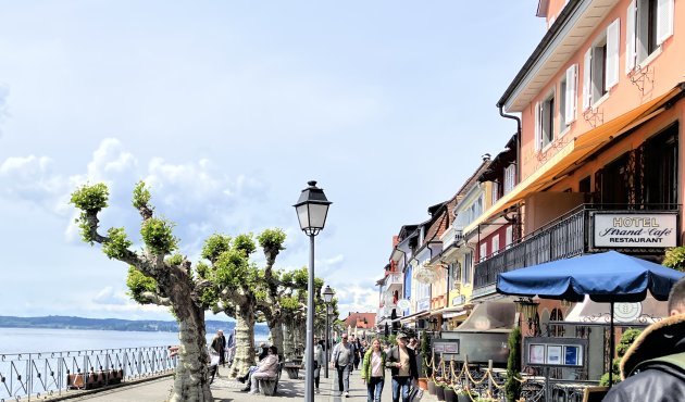 Ein Foto der Strandpromenade in Meersburg am Bodensee. Links der See hinter einem Geländer, davor Bäume und Bänke, flanierende Menschen. Bunte Häuser, Hotels, Cafés und Restaurants