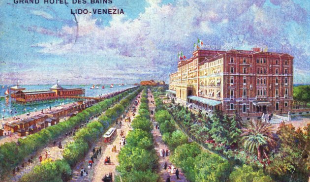 Historische, farbintensive Ansichtskarte vom „Grand Hotel des Bains“ am Lido in Venedig: Rechts steht ein großes, mehrstöckiges Hotelgebäude in warmen Rot- und Ockertönen mit hellen Fensterrahmen und kleinen Flaggen auf dem Dach. In der Bildmitte führt eine breite, von dichtem Grün gesäumte Allee perspektivisch in die Ferne; 