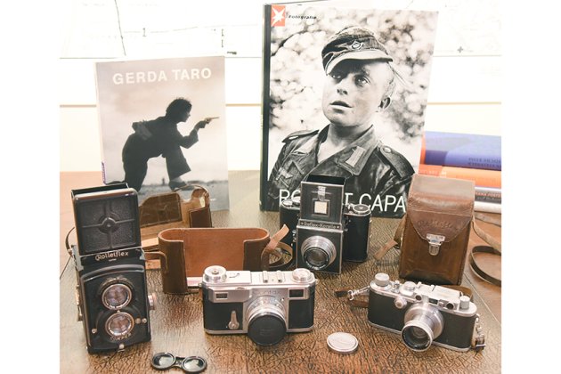 Ein Foto zeigt mehrere Kameras auf einem Schreibtisch, darunter eine Rollei, eine Contax und eine Reflex-Korelle. Im Hintergrund stehen zwei Fotobücher, eines über Gerda Taro und eines über Robert Capa. Die Kameras sind in verschiedenen Größen und Designs angeordnet, und einige sind in Lederetuis verpackt. Der Tisch hat eine strukturierte Oberfläche, und die Umgebung ist hell beleuchtet.