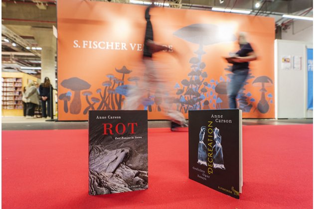 Zwei aufrecht stehende Bücher von Anne Carson stehen auf einem roten Teppich in einer Messehalle. Links ist das Buch „ROT“ mit schwarzem Cover und rotem Titel zu sehen, rechts „DECREATION“ mit dunklem Cover, gelber Schrift und einer blauen Figur. Dahinter hängt eine große orangefarbene Wand mit Pilzillustrationen und der Aufschrift „S. FISCHER VERLAGE“. Vorbeigehende Menschen sind durch die Bewegung unscharf dargestellt.