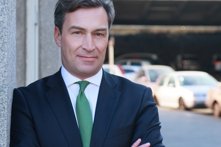 Holger Krimmer in Anzug und grüner Krawatte an eine Häuserwand gelehnt, unscharf im Hintergrund Autos