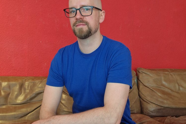 Robin Schmerer mit Brille und blauem T-Shirt auf einer Ledercouch vor einer roten Wand