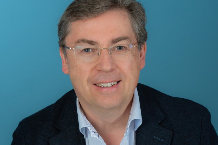 Norbert Gertsch mit Hemd und Sakko, lächelnd vor blauer Fotoleinwand