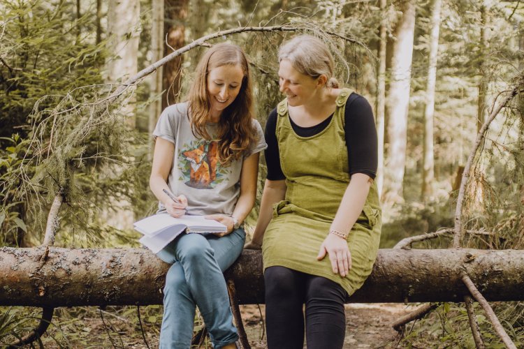 Charlotte Stiefel und Sarah Roller sitzen im Wald auf einem Baumstamm