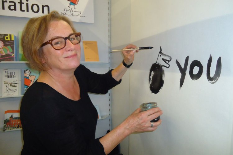 Illustratorin Jutta Bauer hat auf der FBM eine Figur auf die Wand eines Messestands gezeichnet und lächelt in die Kamera