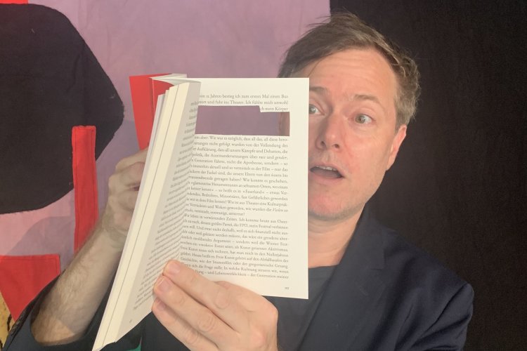 Milo Rau zeigt auf dem Foto ein Buch mit der herausgeschnittenen Stelle