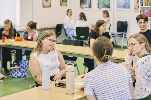 Mehrere junge Erwachsene sitzen in einem hellen Seminarraum an zusammengestellten Tischen und arbeiten in kleinen Gruppen; im Vordergrund diskutieren drei Personen konzentriert miteinander. Auf den Tischen stehen Trinkflaschen und Notizen, im Hintergrund sind weitere Gesprächsrunden und an der Wand farbige Bilder/Plakate zu sehen.