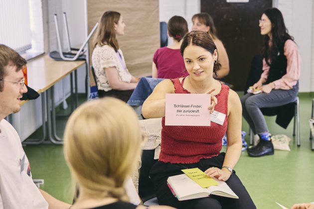 In einem Seminarraum sitzt eine Gruppe junger Erwachsener im Stuhlkreis. Im Vordergrund hält eine Frau in rotem, ärmellosem Oberteil eine rosa Karte hoch; darauf steht „Schlüssel-Finder, der zurückruft (Du rufst an, Schlüssel klingeln)“. 