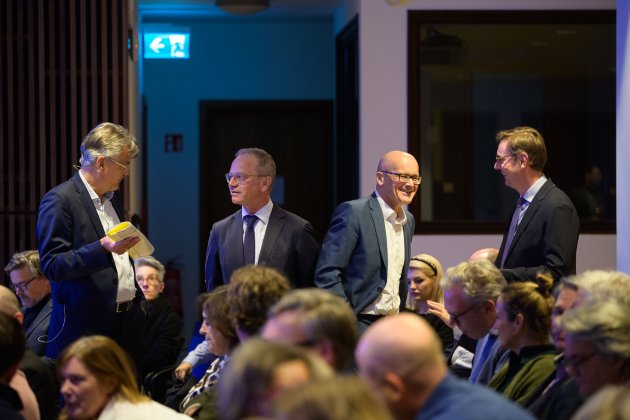 Zwischen sitzendem Publikum stehen 4 Männer, jeweils 2 unterhalten sich miteindner. Namen wir in der Bildbeschreibung