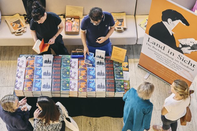 Vogelperspektive auf eine Buchpräsentation. Zwei Personen stehen hinter einem Tisch und blättern in Büchern; davor und darum herum stehen mehrere Besucher:innen. Rechts lehnt ein orangefarbenes Plakat mit der Aufschrift „Land in Sicht – Die Buchhandlung im Nordend“