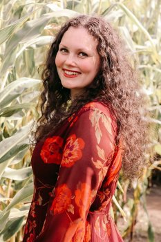 Berenike Woy mit langen lockigen Haaren im Kleid vor unscharfem Hintergrund in einem Getreidefeld