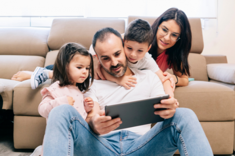 Vater, Mutter und ein Mädchen und ein Junge schauen auf ein Tablet-PC und sitzen vor ihrer Couch