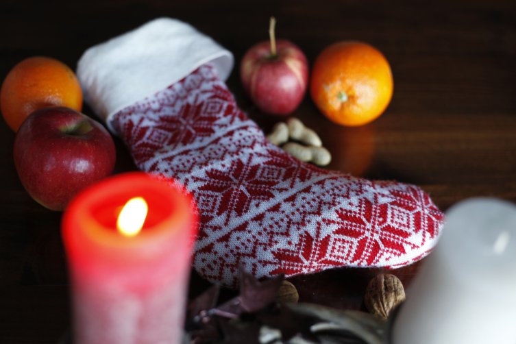 ein im Noprwegermuster gestrickter Nikolausstiefel liegt neben einer brennenden Adventskerze und einem Apfel